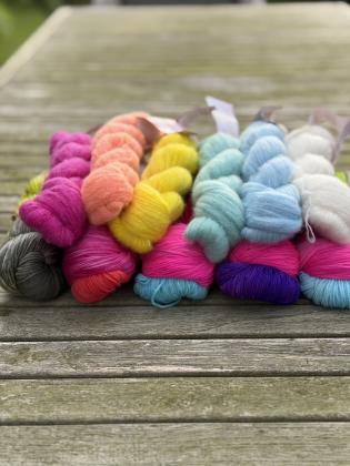 Mehrere handgefärbte Garnkombinationen für das Tuchdesign Petit Provence von Herzfasern auf einem Holztisch im Freien fotografiert. Zu sehen sind verschiedene Farben der Herzfasern No. 11 sowie die fluffige Kaschmir-Seidenqualität Herzfasern No. 93 in unt